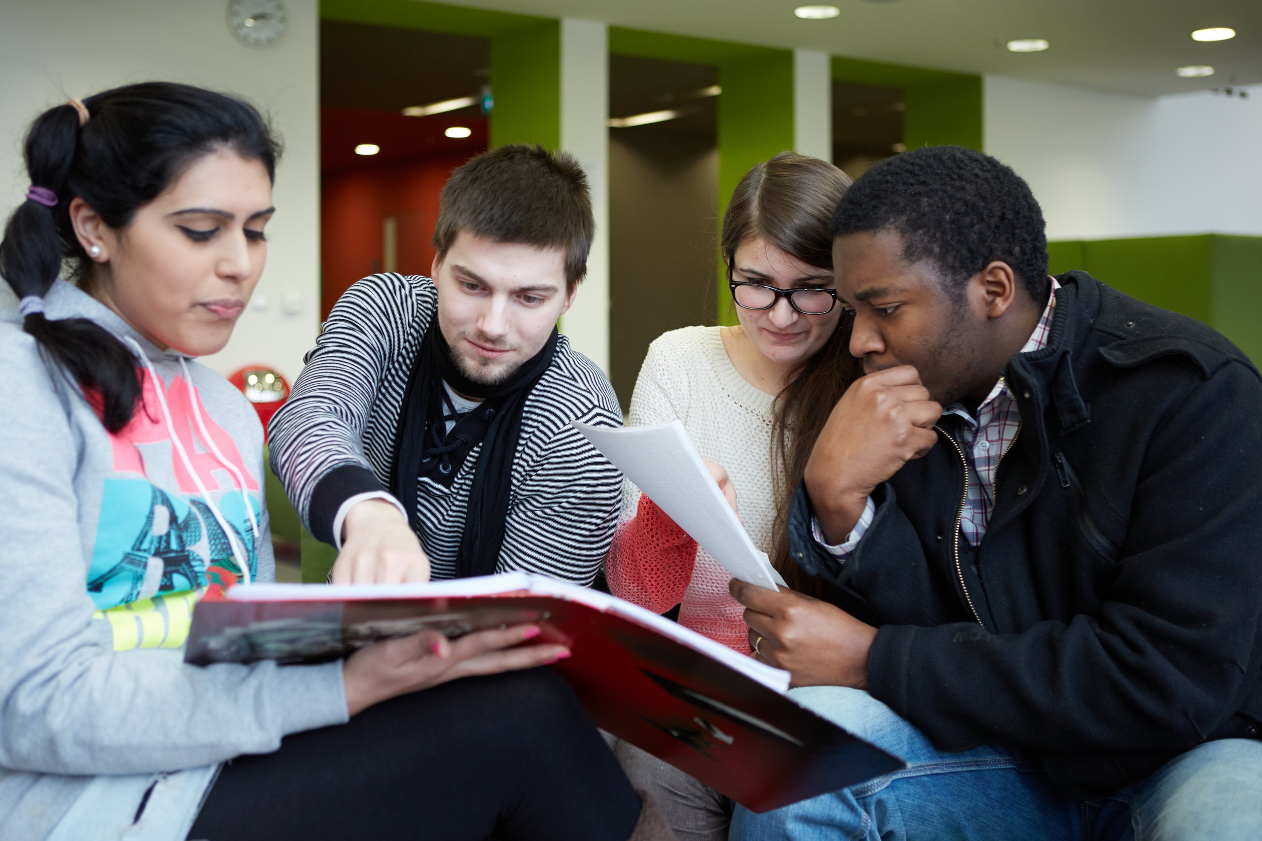 A student shows another student something in an opened book, a third student is sitting next to them