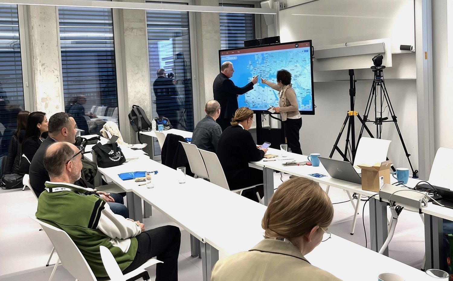 Zwei Personen zeigen einer Gruppe anderer Personen auf dem Smartboard auf der Weltkarte, von welcher Hochschule sie kommen.